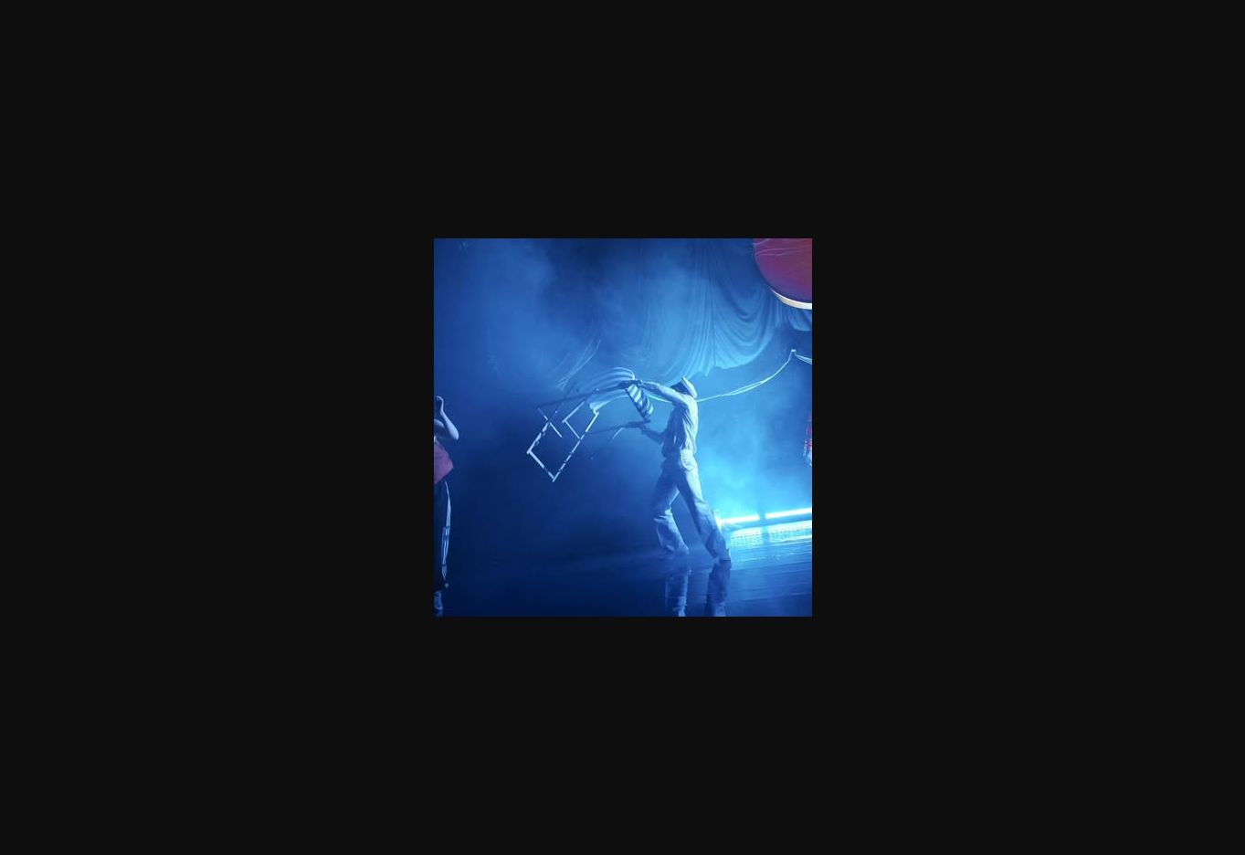 A performer carrying a chair through deep blue stage light